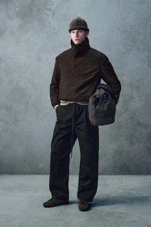 Male model in a dark brown jacket, beanie, and trousers holding a duffel bag against a textured grey background.