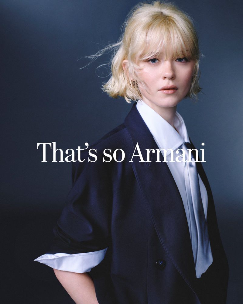 Portrait of a young woman with blonde hair wearing a navy blazer and tie, posing in front of a dark grey backdrop with Armani text.