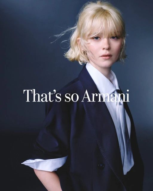 Portrait of a young woman with blonde hair wearing a navy blazer and tie, posing in front of a dark grey backdrop with Armani text.