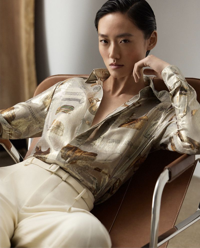 Portrait of an Asian woman in a patterned blouse and white pants, seated in a leather chair against a neutral background.