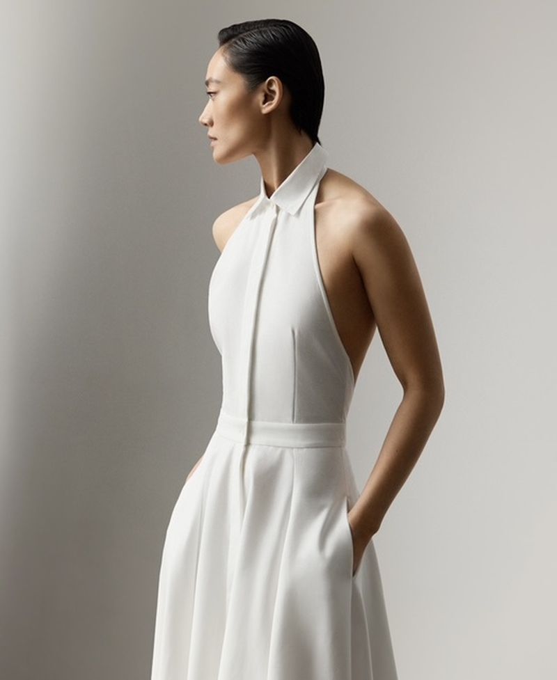 Elegant Asian woman poses in an open-back white halter dress with hands in pockets, side view, in a bright photo studio setting.