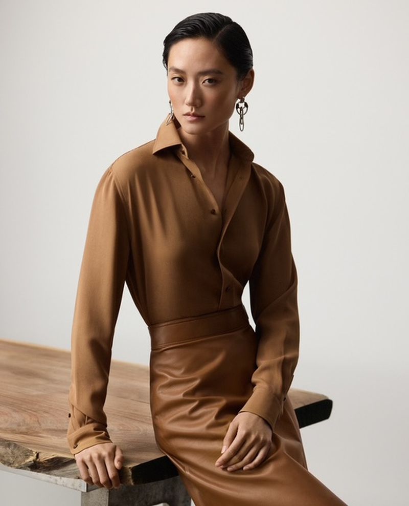 Portrait of a fashion model wearing a brown long-sleeve shirt and matching skirt, leaning on a wooden table in a neutral studio.