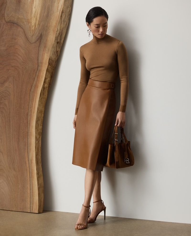 Adult woman poses in a brown top and leather skirt next to a wood panel, accessorized with a brown purse and high-heel shoes.