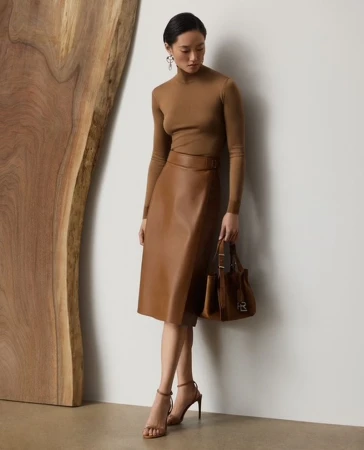 Adult woman poses in a brown top and leather skirt next to a wood panel, accessorized with a brown purse and high-heel shoes.