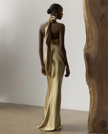Woman in a gold gown with a large bow at the neck posing next to a wooden column in a neutral setting, fashion photography.