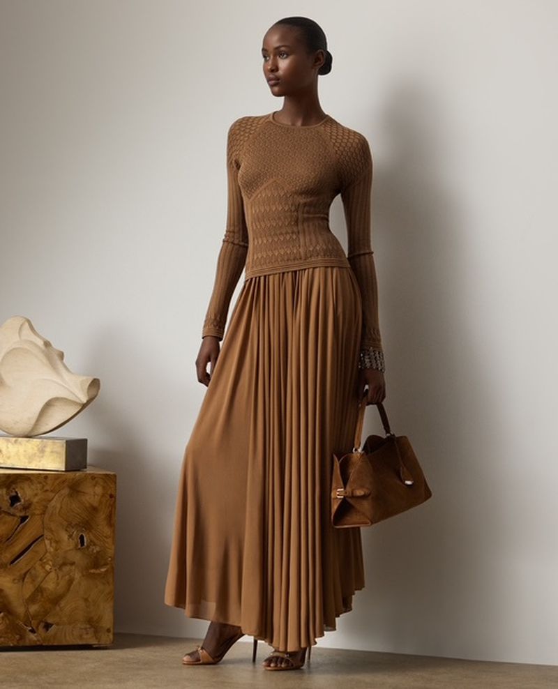 An African American fashion model poses in a long-sleeved knit top and long pleated skirt, holding a handbag against a neutral background.