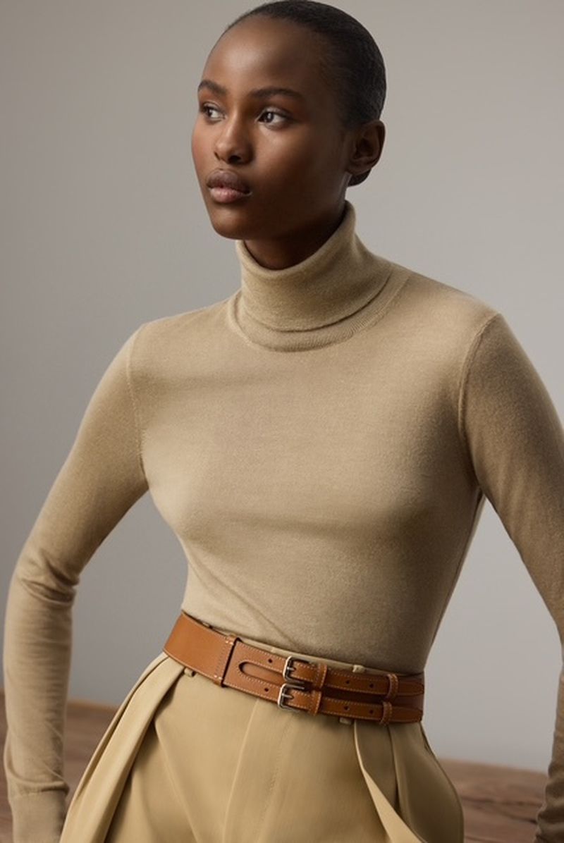 A dark-skinned woman poses in a studio, wearing a turtleneck and skirt with a tan double-buckle belt, highlighted by soft, neutral lighting.