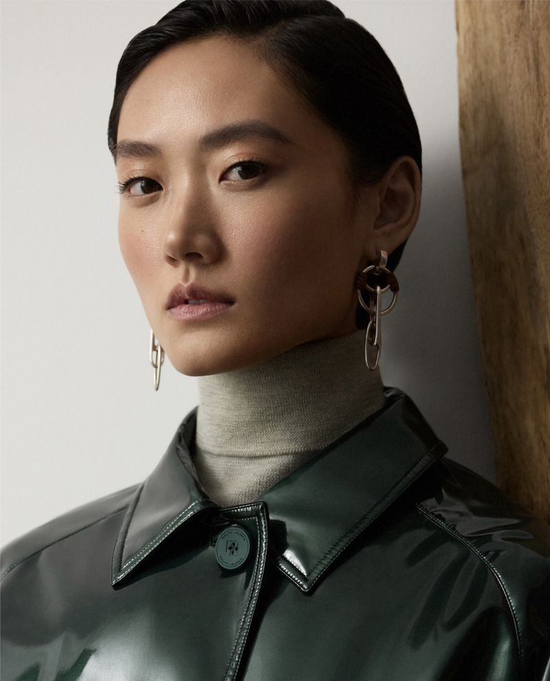 Headshot of a woman with dark hair in a glossy green jacket and a grey turtleneck sweater, wearing dangling earrings in a studio setting.