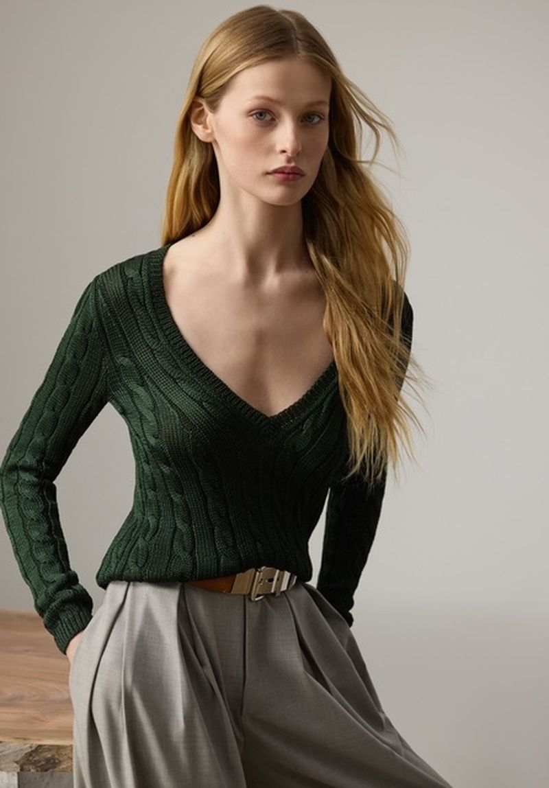 A fair-skinned fashion model poses indoors wearing a green, cable-knit V-neck sweater, tan belt, and pleated, wide-leg grey pants in a studio setting.