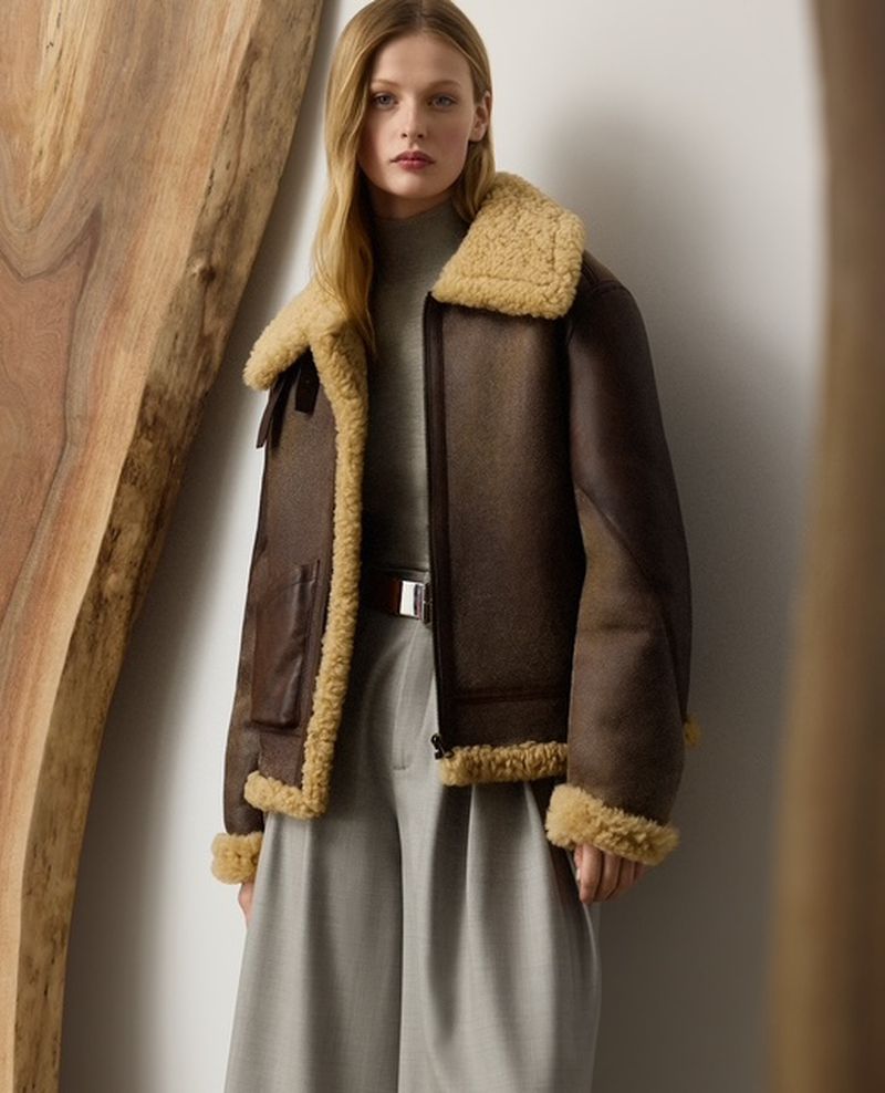 Model wearing a brown shearling-lined aviator jacket, grey turtleneck, and pleated wide-leg trousers standing beside a wood slab.