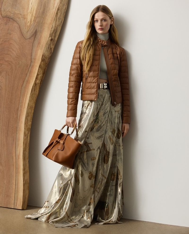A woman in a brown jacket and metallic skirt holding a leather handbag in front of a wood panel and white wall.