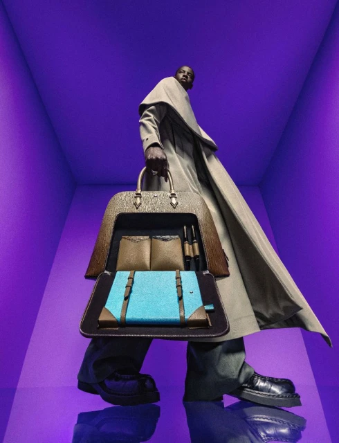 A stylish adult male carries a teal and brown luxury bag with an open compartment against a monochromatic violet studio backdrop.