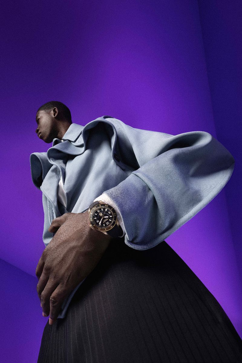 An African American male fashion model poses with a wristwatch against a purple background in a studio, shot from a low angle.