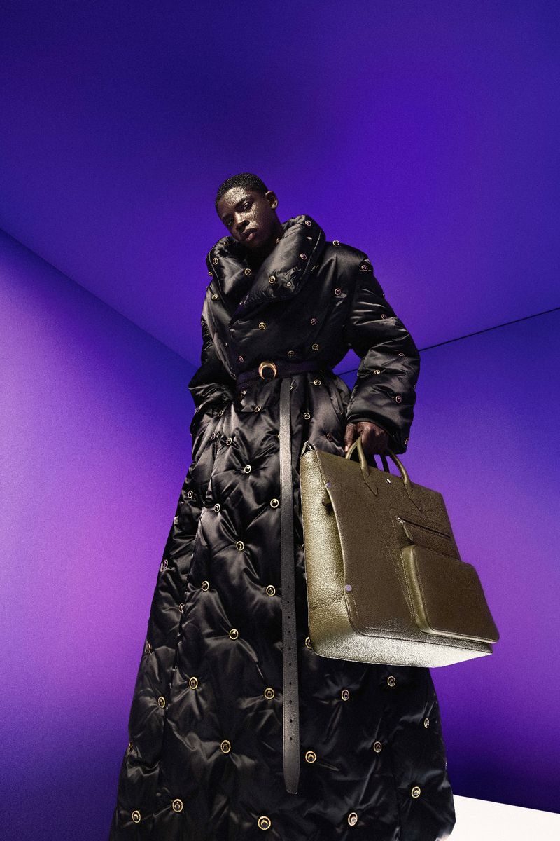 Dark-skinned model wearing a black, quilted, belted coat and carrying a beige bag in a studio with purple-gradient walls.