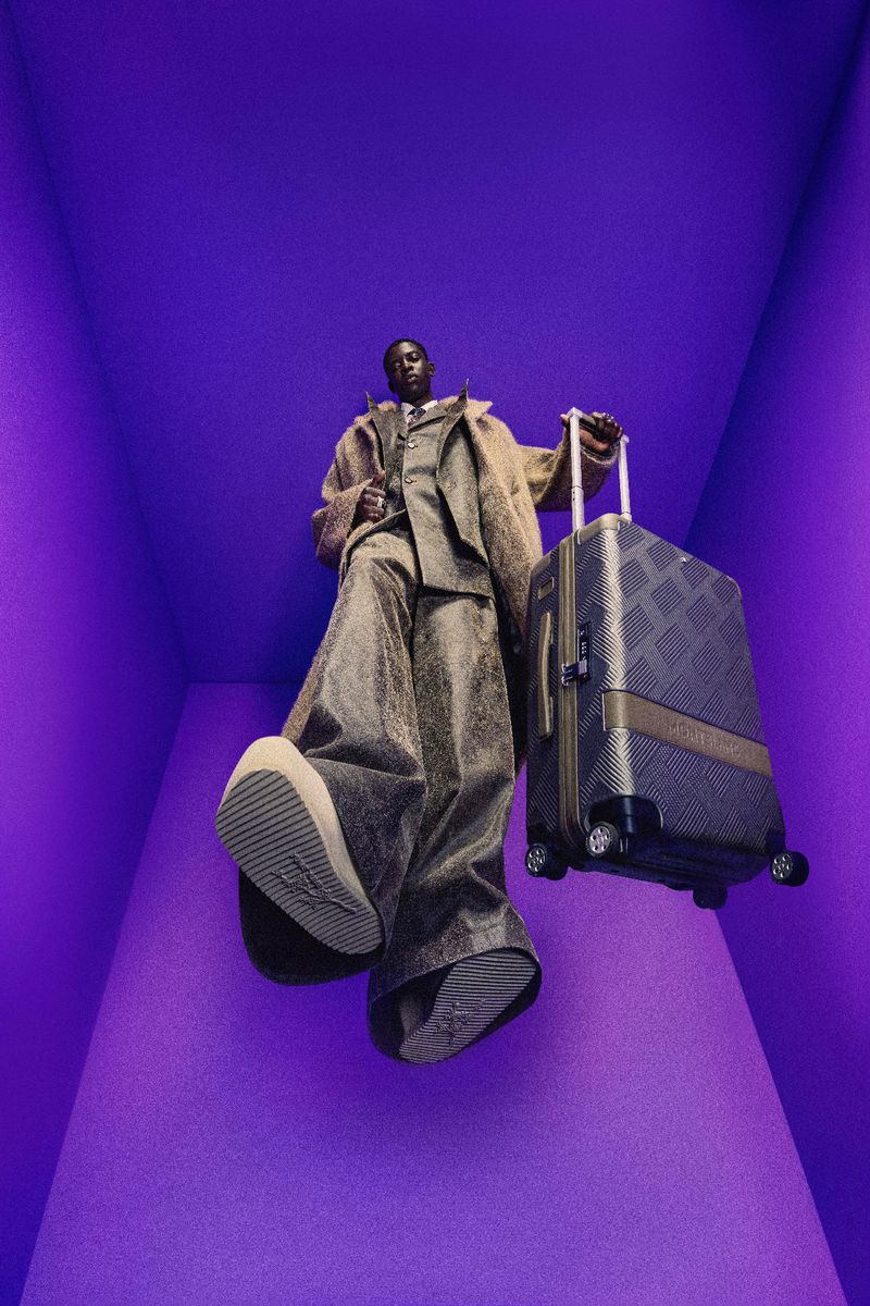 A stylish Black male model poses in a purple room, wearing a brown suit and holding a checkered luggage, photographed from a low angle.