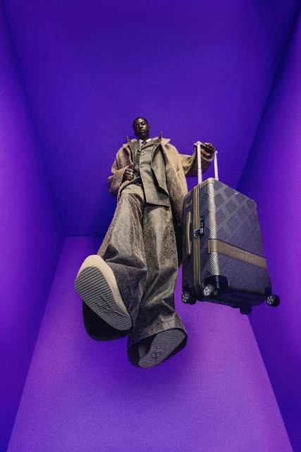A stylish Black male model poses in a purple room, wearing a brown suit and holding a checkered luggage, photographed from a low angle.