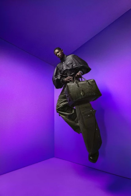 Fashionable Black man strikes a pose in a bright purple studio, showcasing a sophisticated black jacket, olive cargo pants, and leather duffel bag.