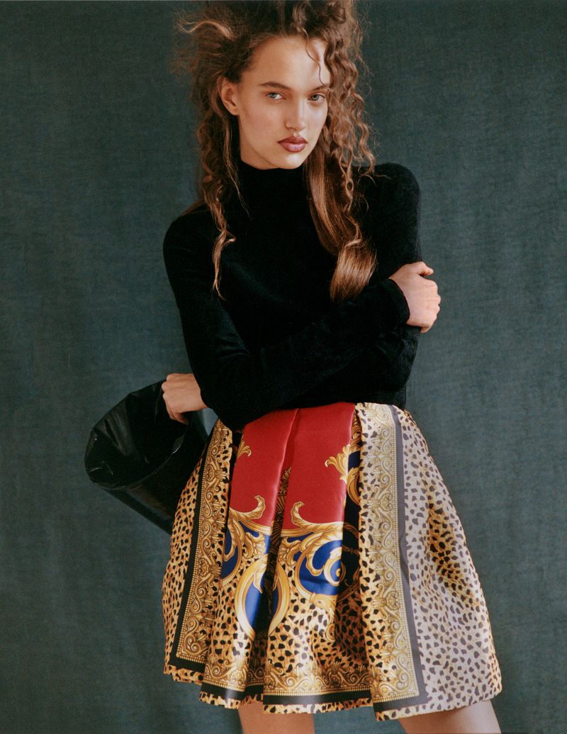 Fashion shot of a model in a black turtleneck and patterned skirt holding a black bag, posing against a muted green backdrop.