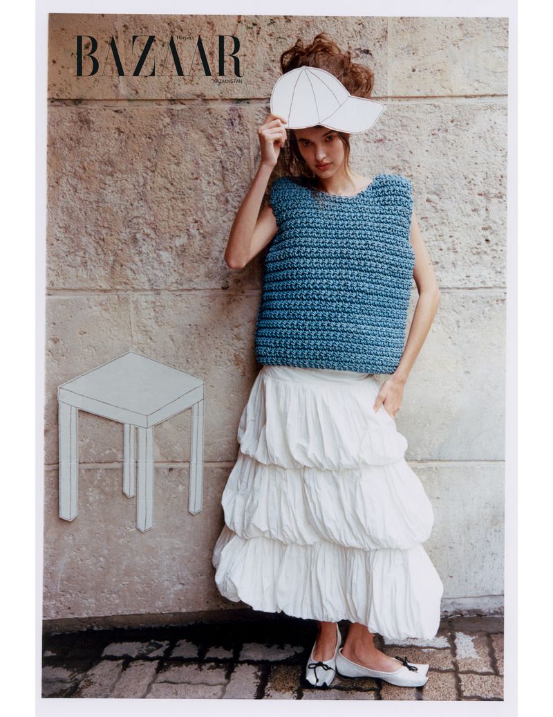 Fashion editorial image featuring a female model in a blue knit vest and a white tiered skirt, against a textured wall with BAZAAR logo.