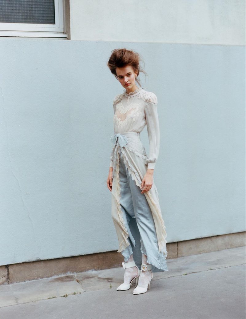 A fashionable adult woman in a sheer top and pantsuit poses in an urban setting against a blue wall, complemented by white ankle boots.