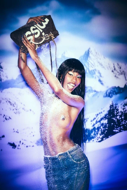 A topless woman in a blue denim mini skirt poses in front of a mountain backdrop, holding a black purse with white letters.