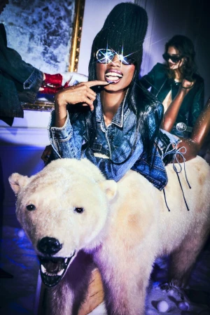 Woman with sunglasses rides a polar bear sculpture indoors with additional people and an ornate gold frame on the wall visible.