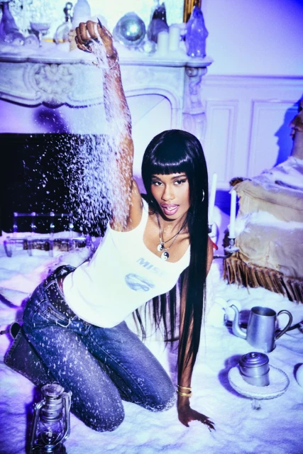 Young woman kneels in a snowy setting, holding and pouring snow. She wears a white tank top and jeans in a staged wintry scene.