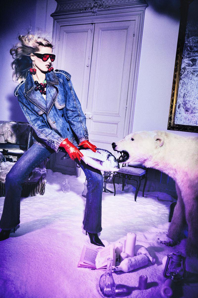 Fashionable adult woman poses with a polar bear statue inside a decorated room with props, wearing a denim jacket and sunglasses.