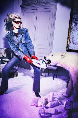 Fashionable adult woman poses with a polar bear statue inside a decorated room with props, wearing a denim jacket and sunglasses.