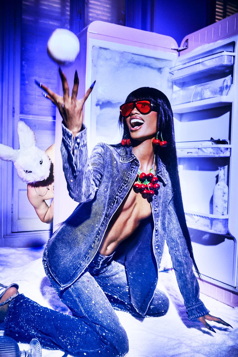 Adult woman poses in denim clothing with red glasses and cherry jewelry, next to a refrigerator and holding a toy rabbit.