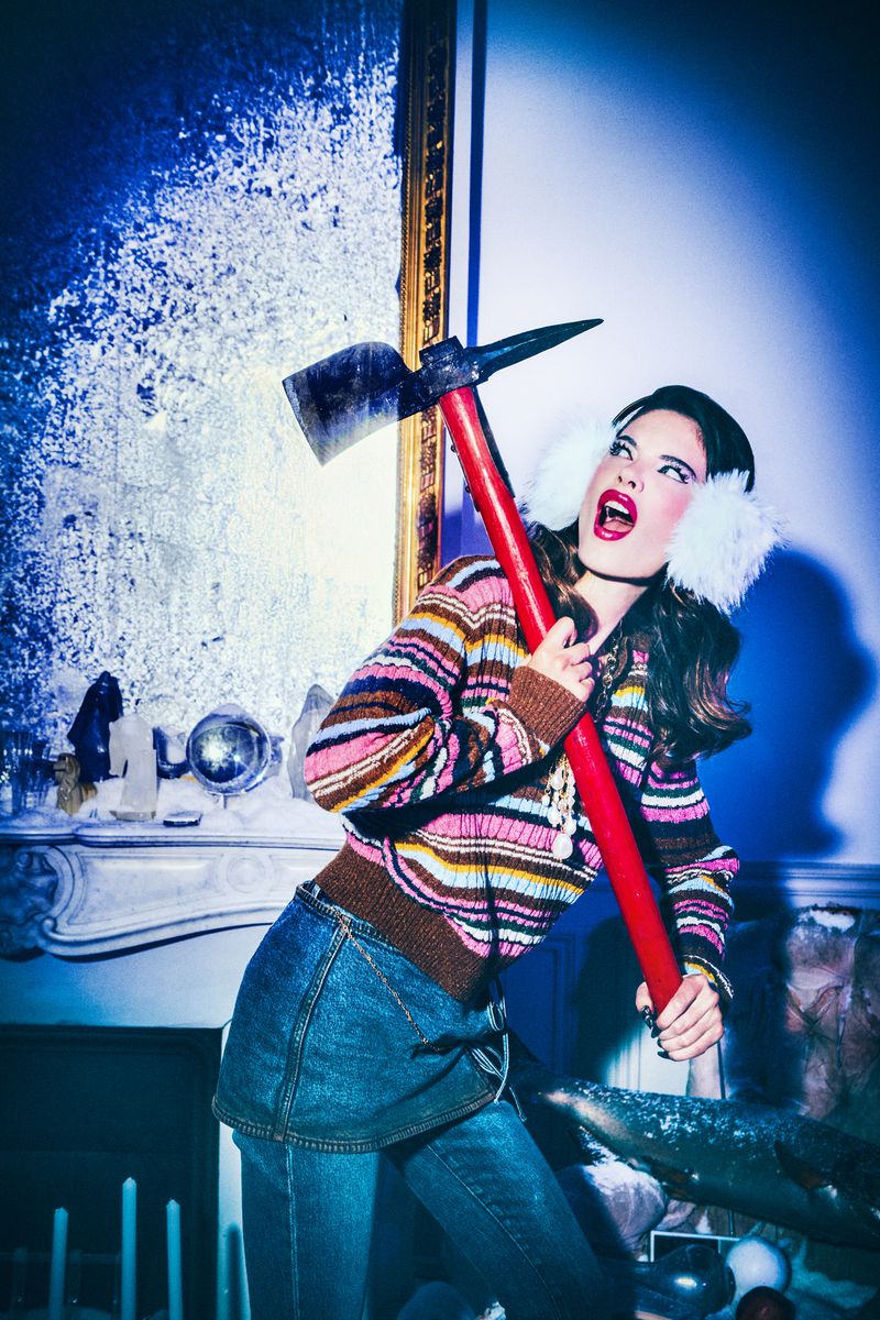 A stylish woman in a striped sweater holds an axe in front of a fancy interior with a decorative mirror and fireplace.