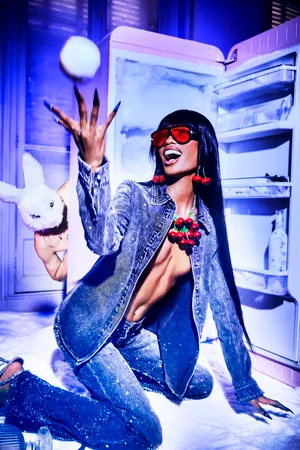 Model in acid-wash denim and red cherry jewelry tosses a snowball in front of an open retro pink freezer with a bunny-masked figure behind.