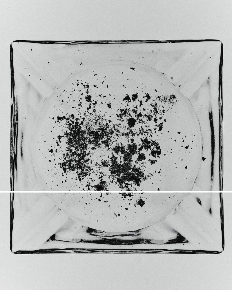 Overhead shot of a clear, square ashtray with black ash scattered inside on a white surface, providing a minimalist, stark aesthetic.