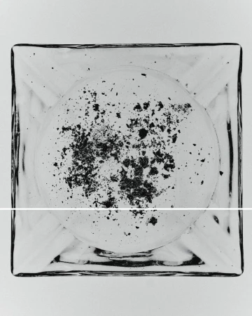 Overhead shot of a clear, square ashtray with black ash scattered inside on a white surface, providing a minimalist, stark aesthetic.