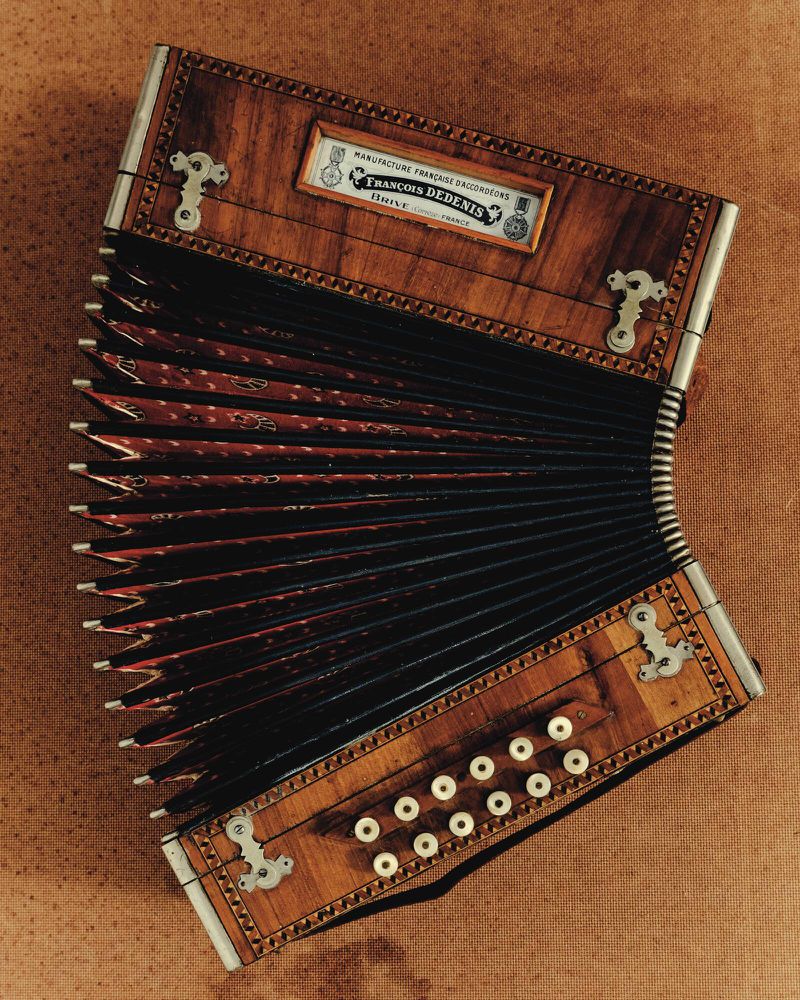 An antique wooden accordion sits on a textured brown surface, featuring intricate marquetry, mother-of-pearl buttons, and ornate metalwork.