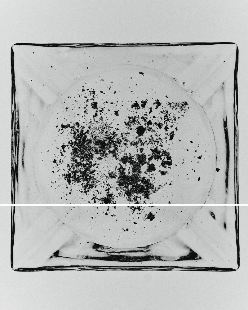 Top-down view of a square clear glass ashtray containing scattered dark ash flakes against a plain white background.