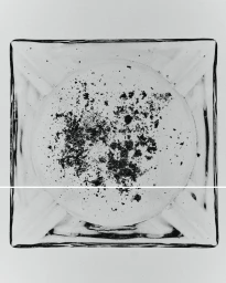 Top-down view of a square clear glass ashtray containing scattered dark ash flakes against a plain white background.