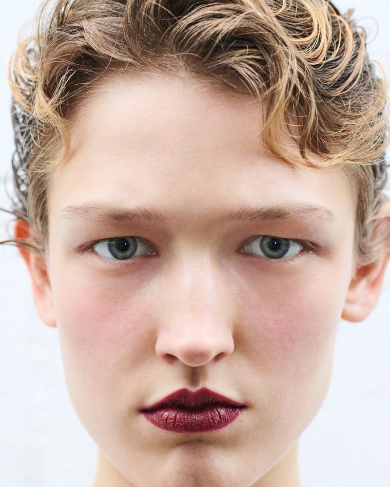 Close-up portrait of a model with short blonde hair and dark red lipstick against a plain white background