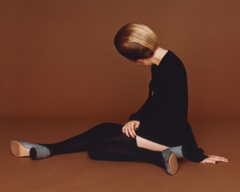 Woman with blonde bob hairstyle poses in black clothing and grey heeled shoes against a brown backdrop in a studio fashion shot.