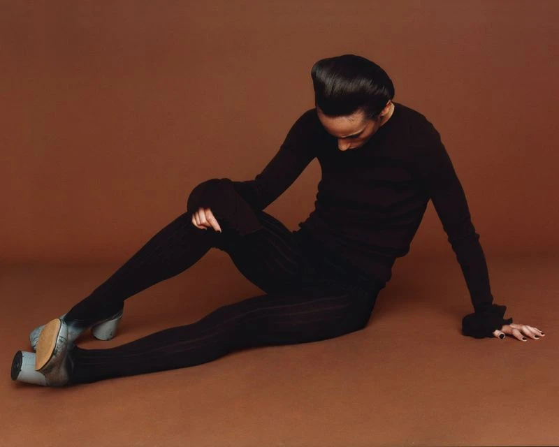 A woman sits on the floor in a black outfit and shoes with sculptural heels, set against a brown backdrop in a studio shot.