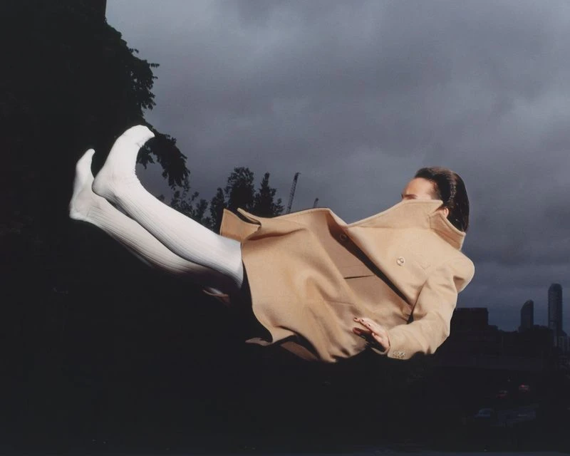 Fashion photography of a woman in a beige trench coat, floating in air against a cloudy sky and a cityscape in the background.