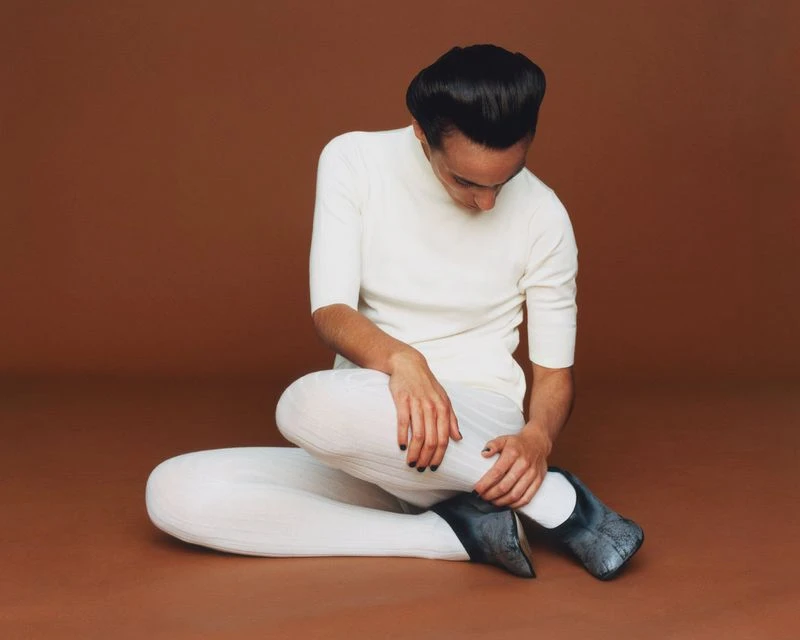 Stylish adult sits cross-legged wearing white top, white ribbed pants and grey shoes against brown backdrop in studio setting.