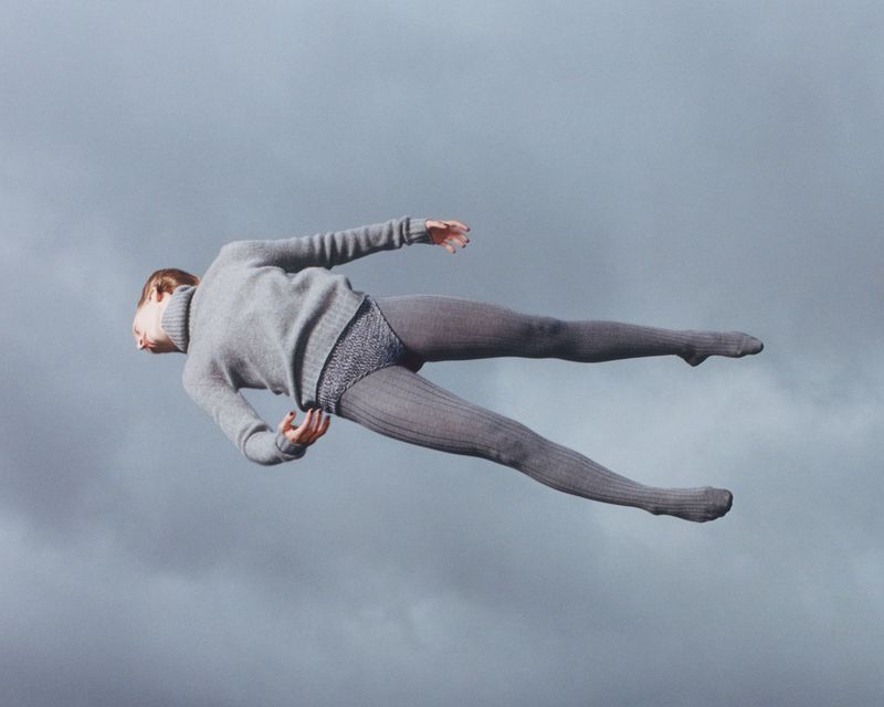 Full shot of a woman floating horizontally against a cloudy sky backdrop, dressed in a grey sweater and tights, creating a surreal effect.