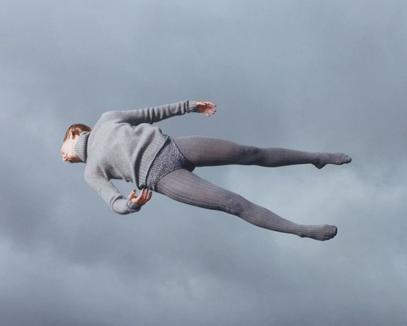 Full shot of a woman floating horizontally against a cloudy sky backdrop, dressed in a grey sweater and tights, creating a surreal effect.