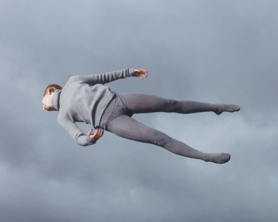 Full shot of a woman floating horizontally against a cloudy sky backdrop, dressed in a grey sweater and tights, creating a surreal effect.