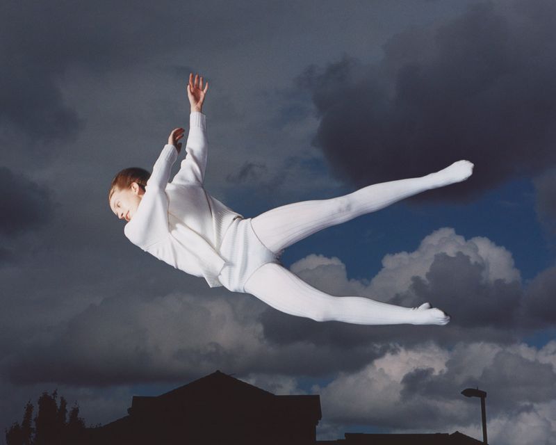 Person in white sweater and pants jumps in front of dark, dramatic clouds with a blurred building in the background, viewed at eye-level.