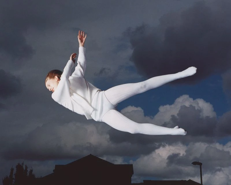 Person in white sweater and pants jumps in front of dark, dramatic clouds with a blurred building in the background, viewed at eye-level.