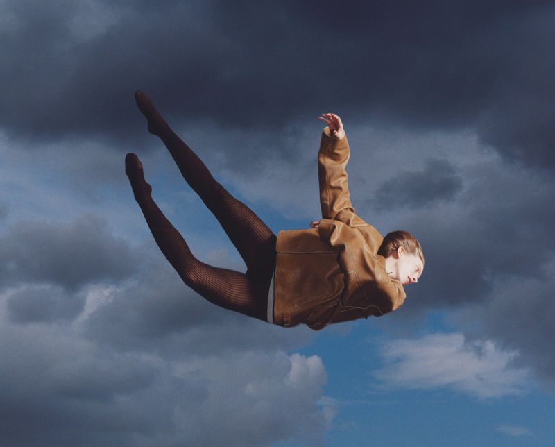 Woman in brown jacket and patterned tights appears to float against a cloudy sky, with soft lighting and a unique pose.