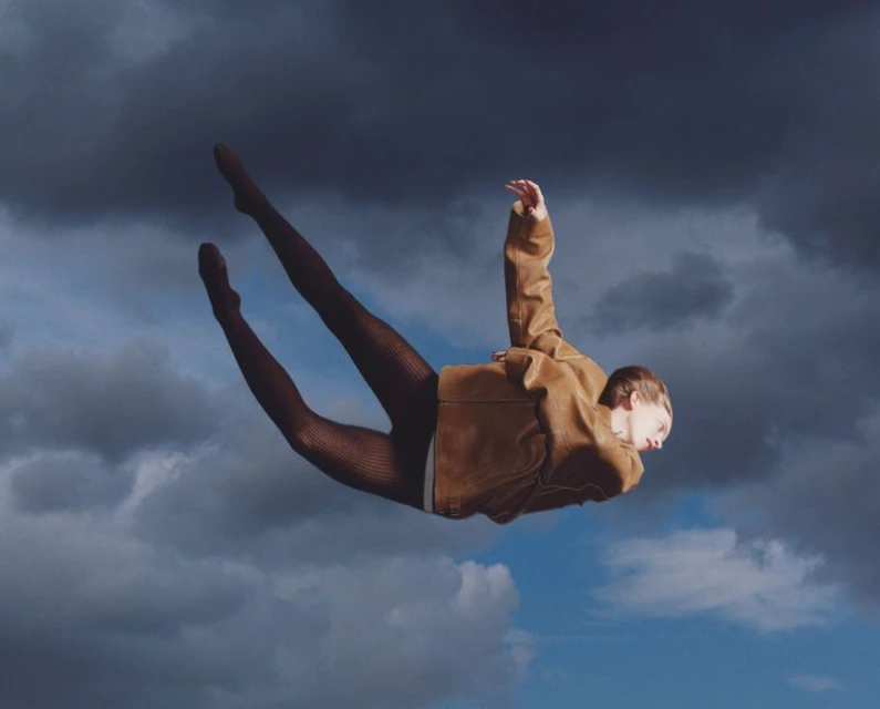 Woman in brown jacket and patterned tights appears to float against a cloudy sky, with soft lighting and a unique pose.