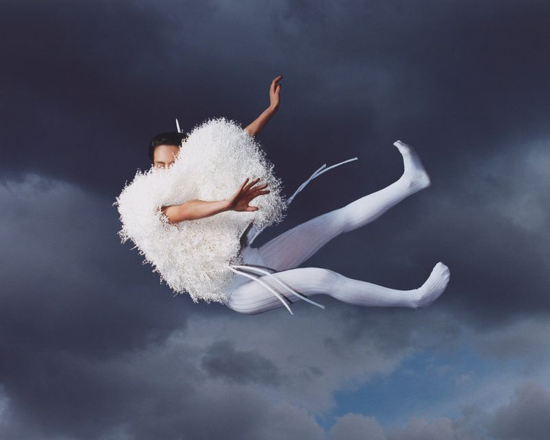 Person in white costume levitating against a stormy sky. The outfit features fluffy shoulders and textured leggings, creating an ethereal effect.